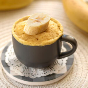 Photo of mug cake in gray mug, with slice of banana sitting atop cake.