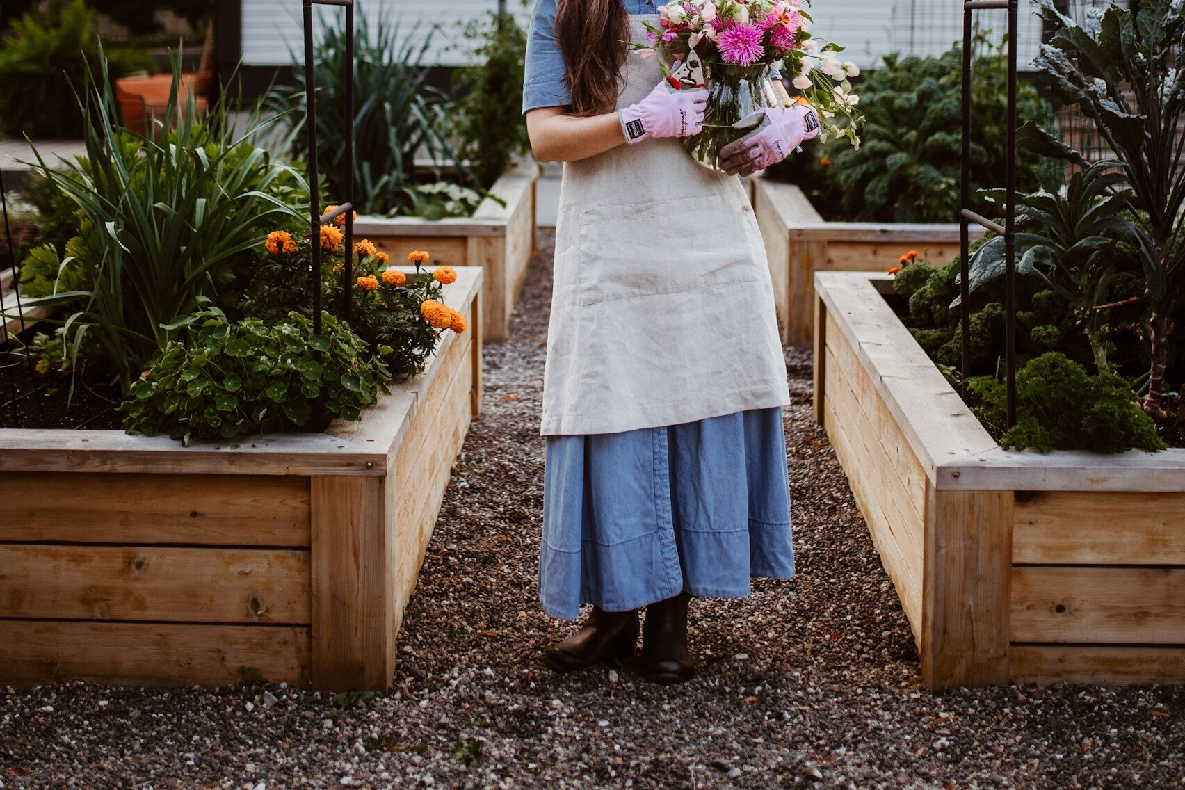 Woman in a denim dress and linen apron standing in her backyard garden that's made of cedar raised beds.