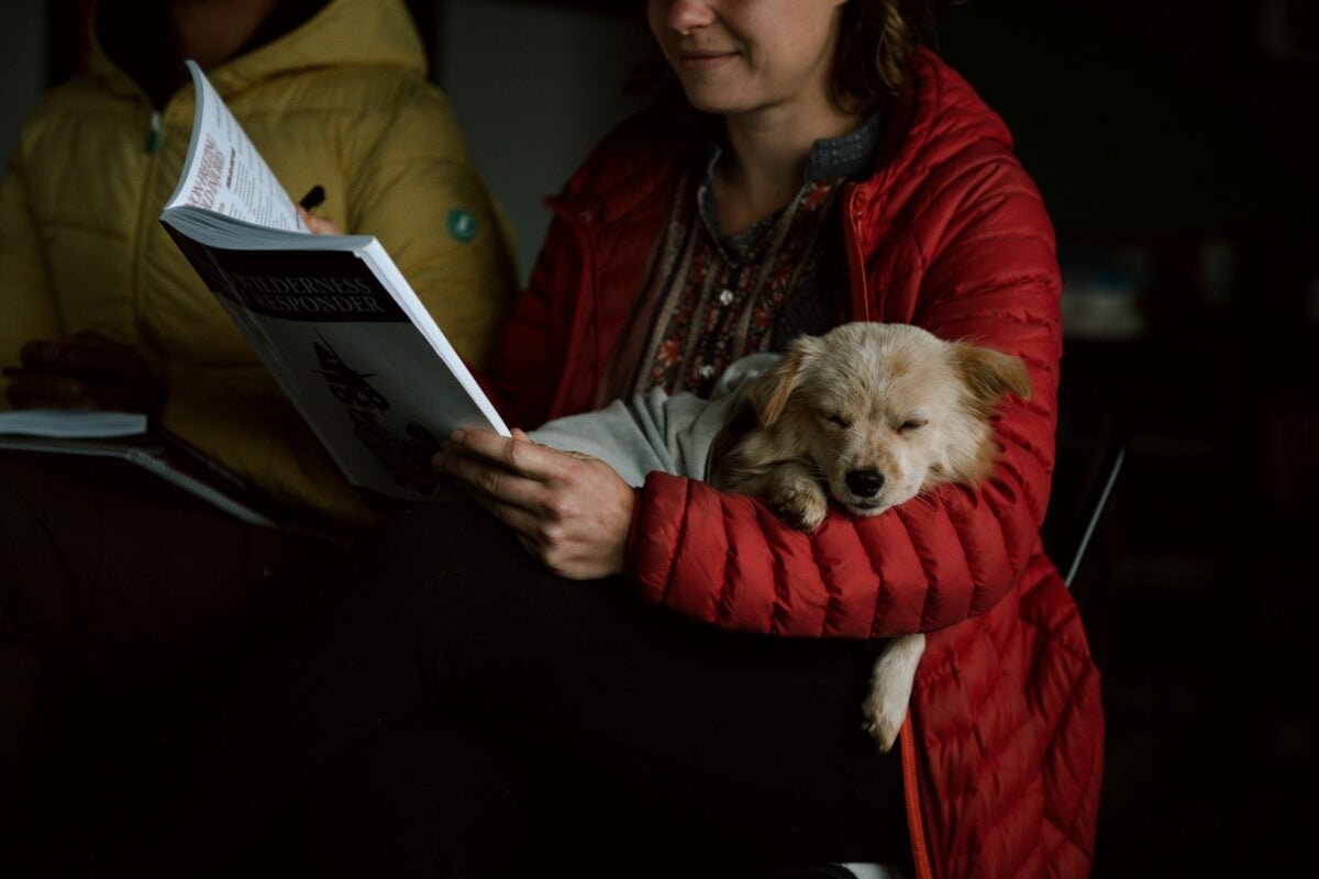 A woman reading while a dog sleeps in her lap.
