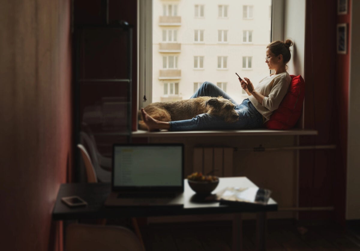 Young woman freelancer sitting at home with dog looking into mobile phone screen.