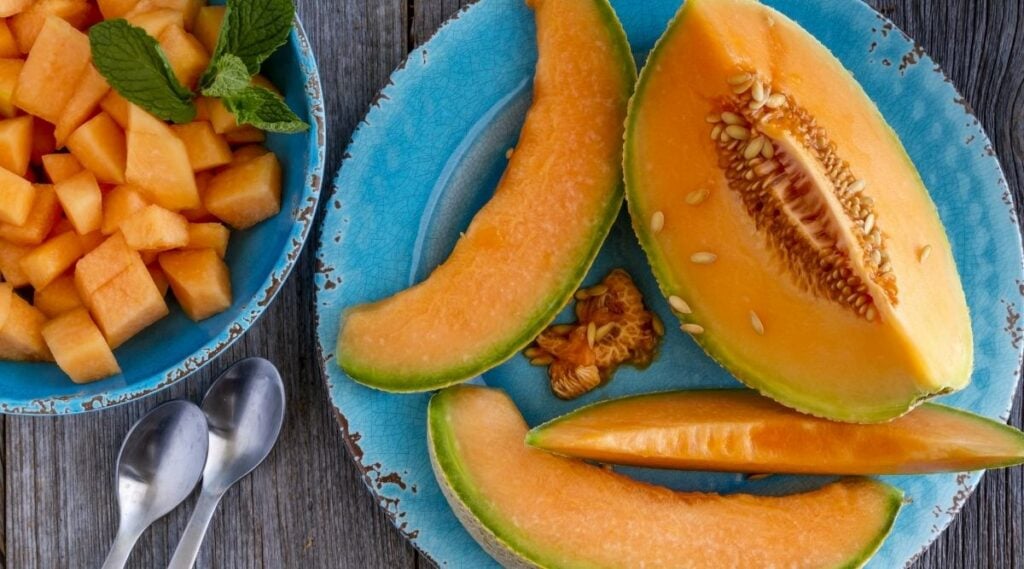 Cantaloupe melon slices sitting on blue rustic plate on wooden tabletop with two spoons and bowl filled with melon pieces
