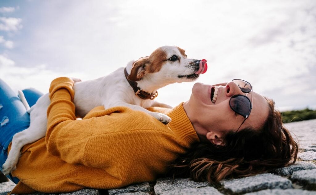 Dog licking a girl's face lying on her back outside.