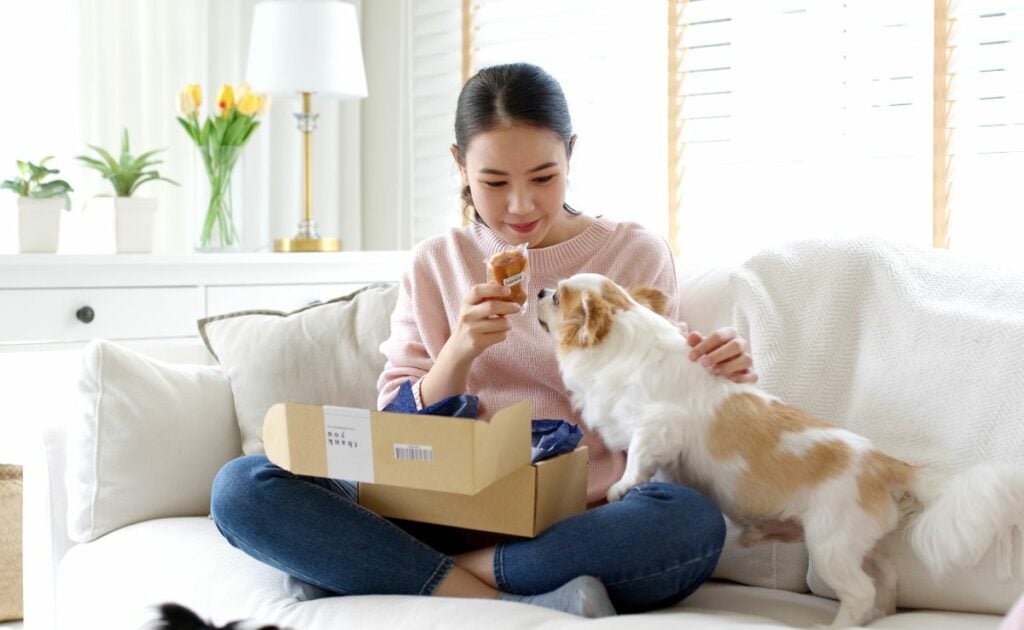 Girl sitting on sofa at home in living room unboxing dog food and snacks with dog in her lap.