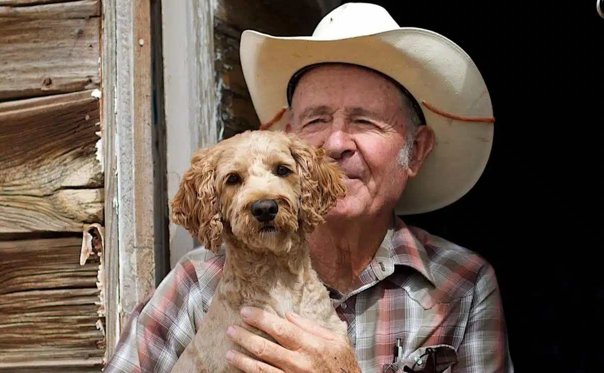 old man with a cowboy hat on holding a dog on a texas ranch