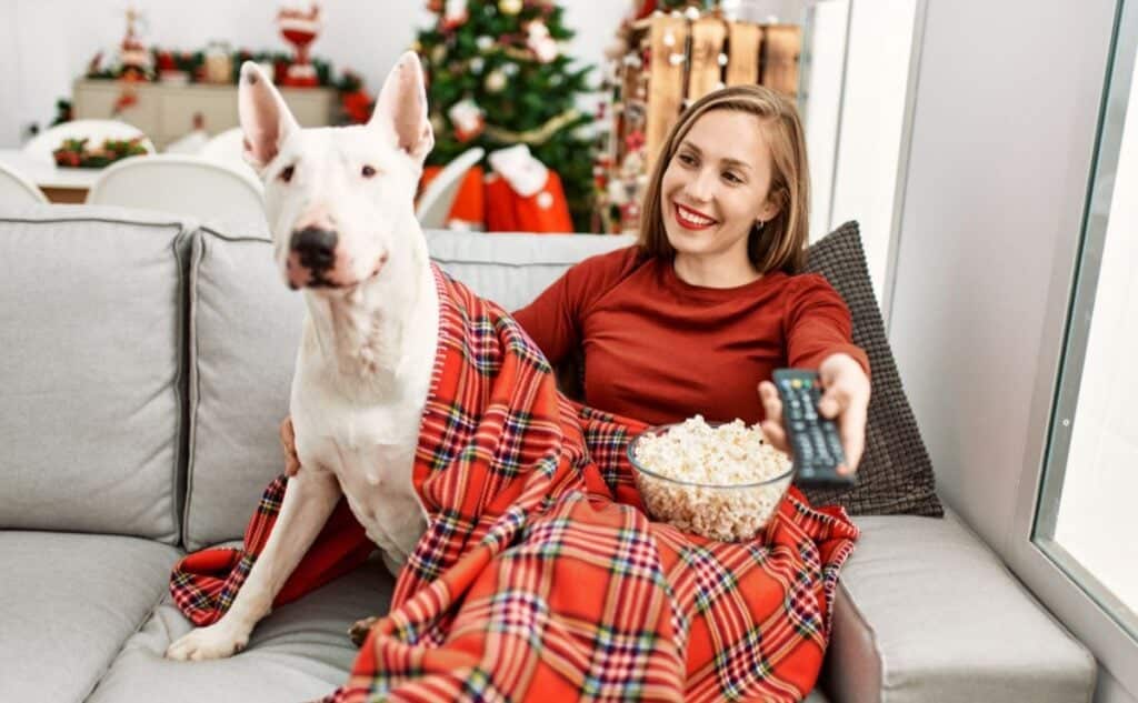 Woman curled up with dog on sofa in Christmas PJs watching movie.